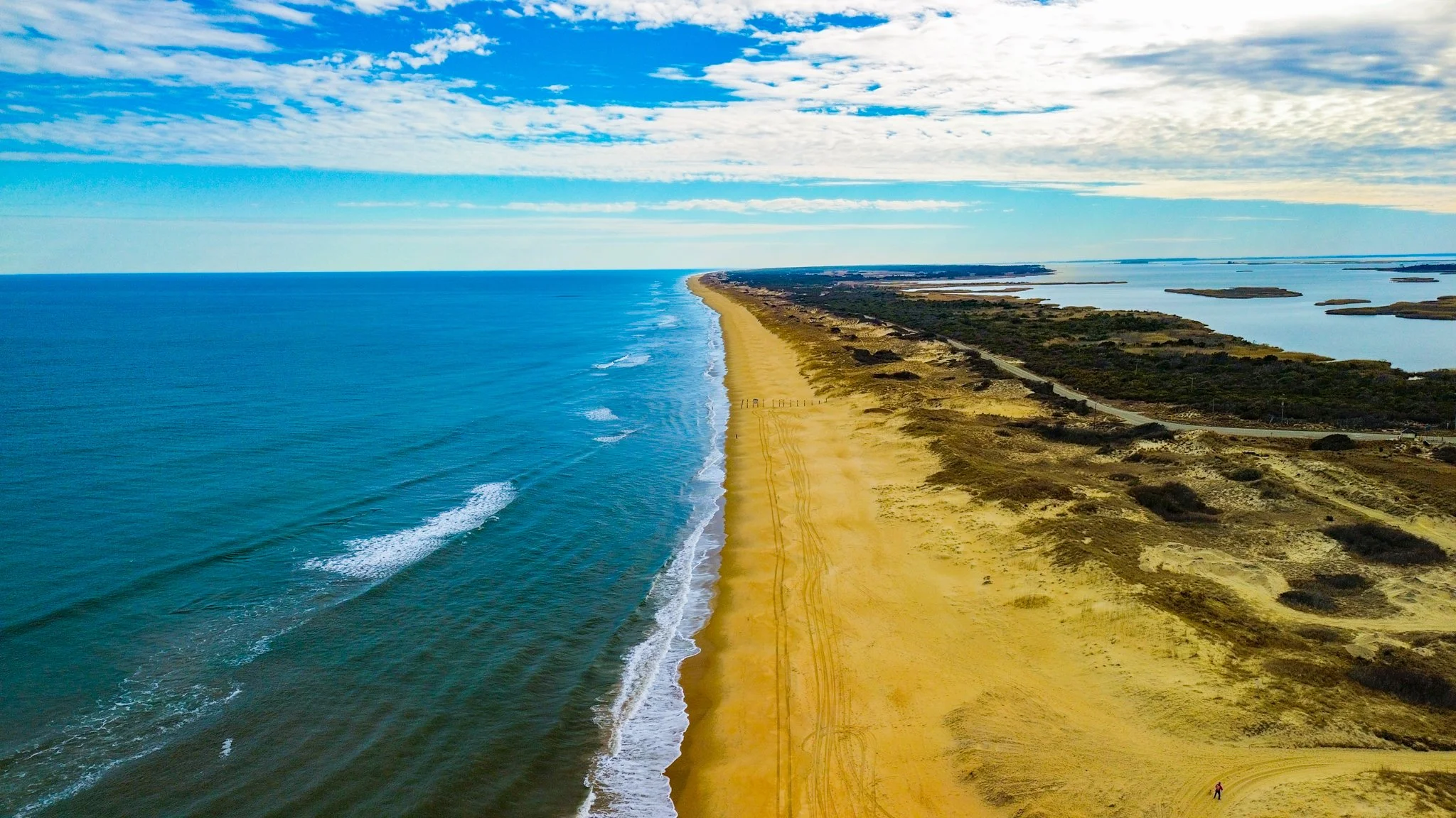 Aerial view of a long sandy beach with waves on the left and dunes on the right, stretching along the coast under a partly cloudy sky.