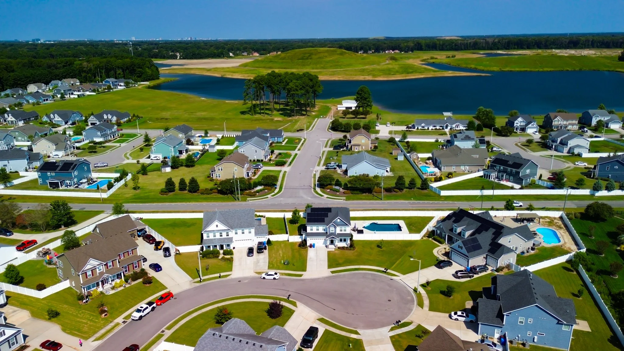 Aerial view of a suburban neighborhood with houses, lawns, roads, pools, trees, and a lake in the background.