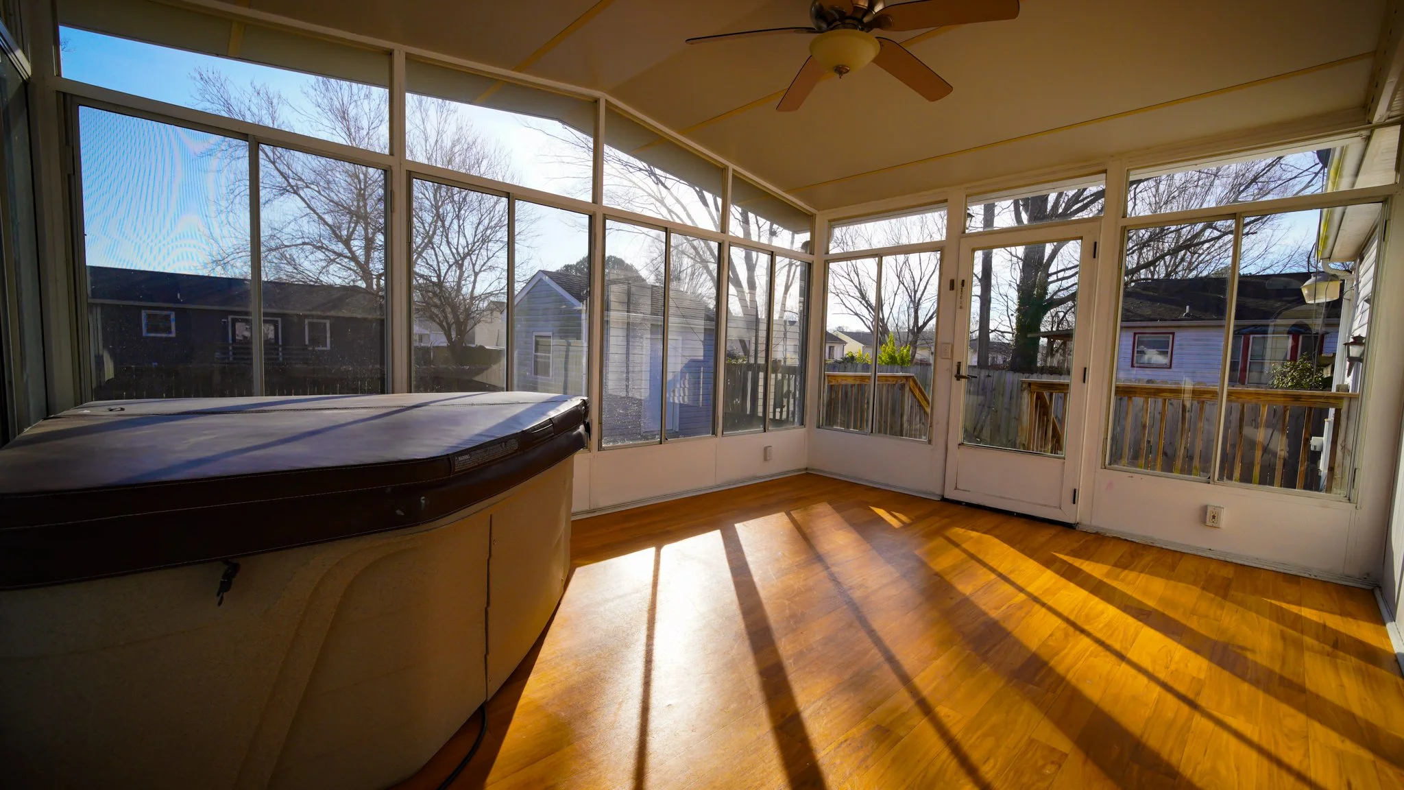 Sunlit enclosed porch with wood floor, large windows, a ceiling fan, and a hot tub in the corner, overlooking neighboring houses and trees.