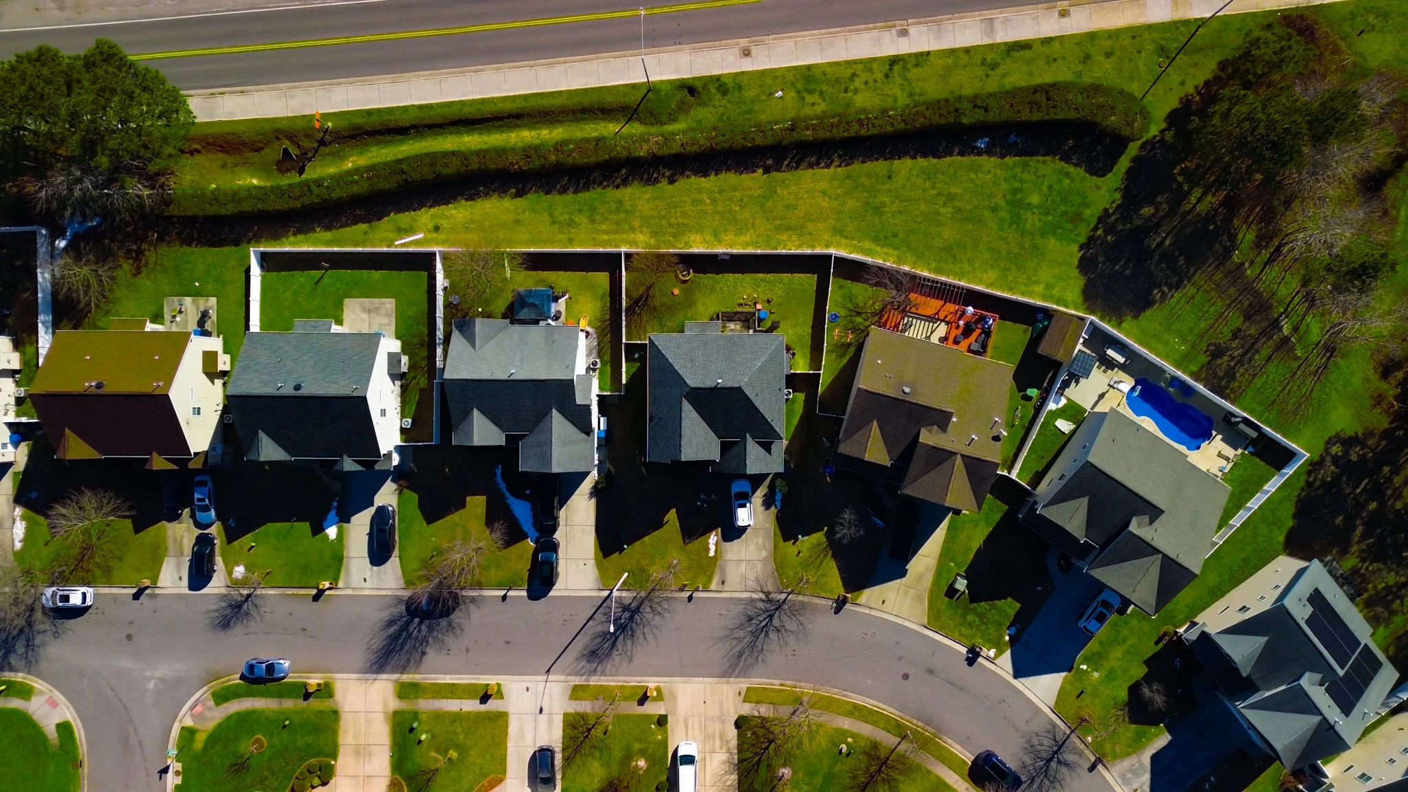 Aerial view of a neighborhood showing houses, driveways, cars, a street, and a backyard with a swimming pool and outdoor deck.