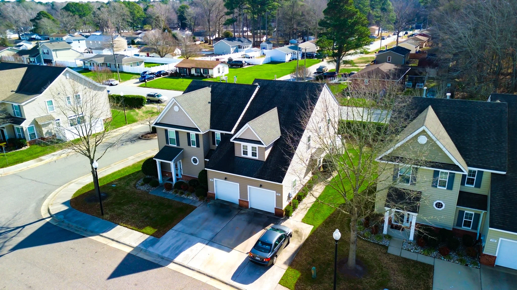 An aerial view of a suburban neighborhood showing multiple detached houses, trees, green lawns, curving streets, and parked cars on driveways and the street.