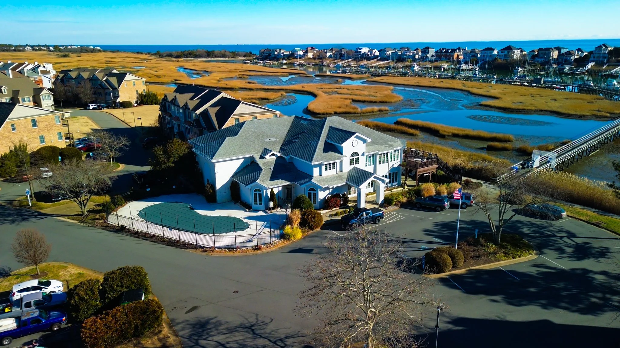 An aerial view of a large white house with a gray roof, parked cars, trees, and a parking lot, overlooking a marshland and waterfront community.