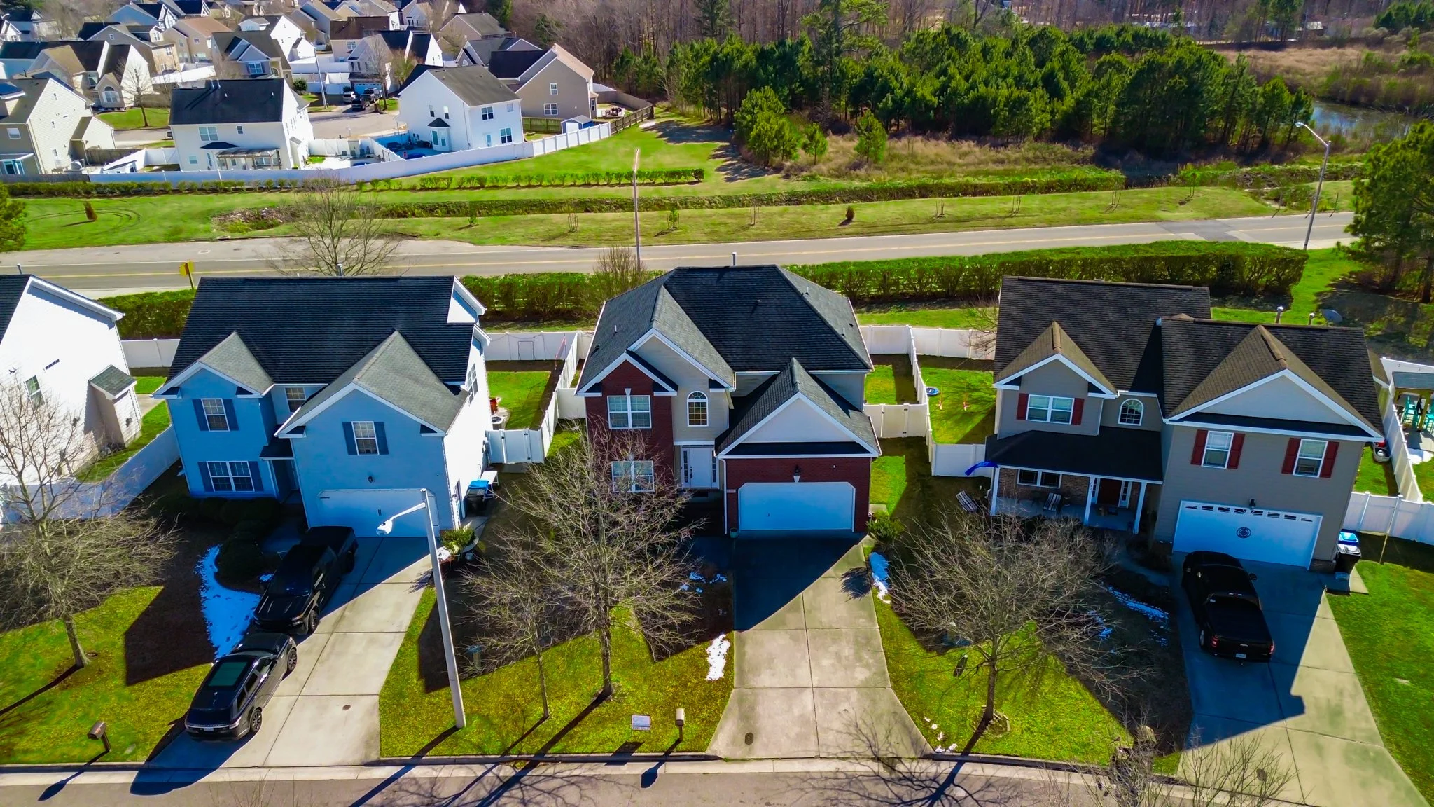 Aerial view of three suburban houses with driveways, parked cars, trees, yards, and a street in front, with a wooded area and a road in the background.