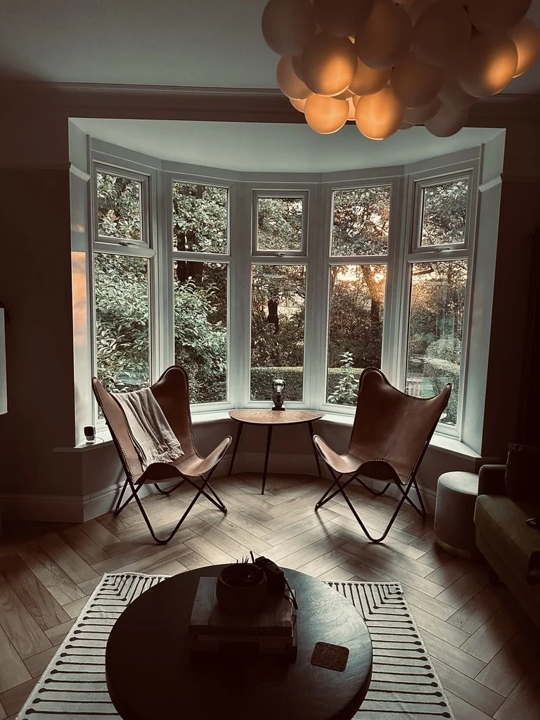 A living room corner featuring two brown leather butterfly chairs and a small round wooden table in front of large bay windows revealing a lush green outdoor view at sunset. There is a white-striped rug on the wooden floor, and a round coffee table with books, a small decorative object, and a coaster is visible in the foreground. A modern ceiling light fixture with spherical bulbs is hanging overhead.