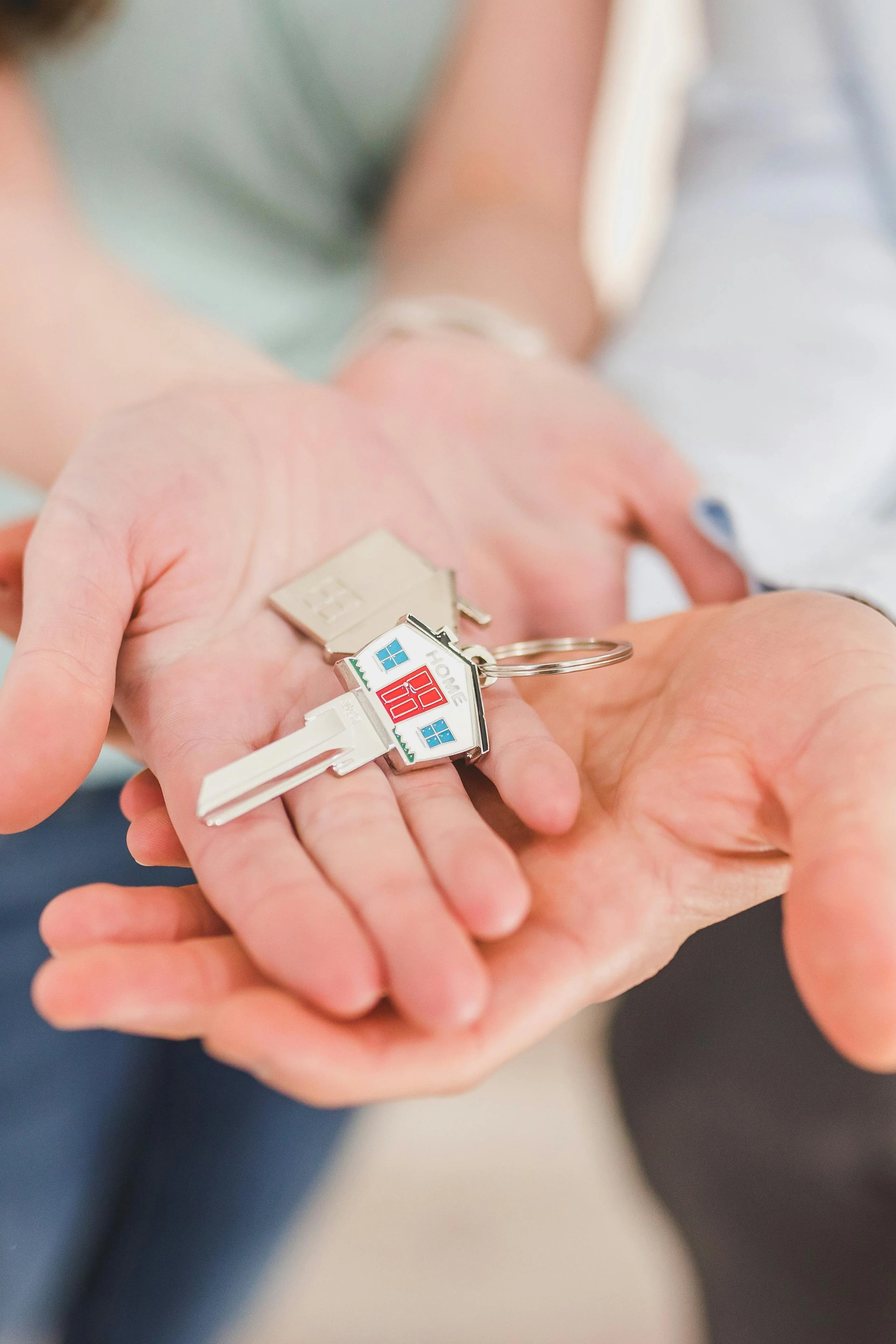 A person's hand holding a set of house-shaped keychains, including a real key, a house photo keychain, and a house-shaped keychain with 'HOME' written on it.