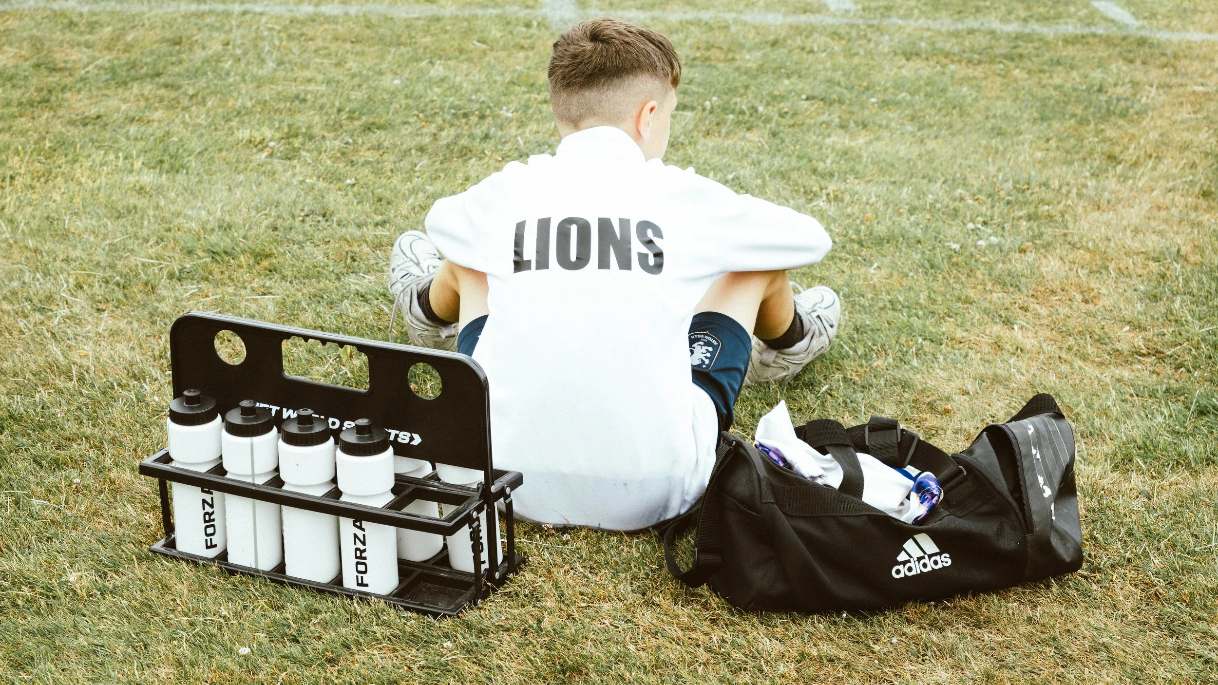A young soccer player sitting on grass field with a water bottle holder and a sports bag nearby.