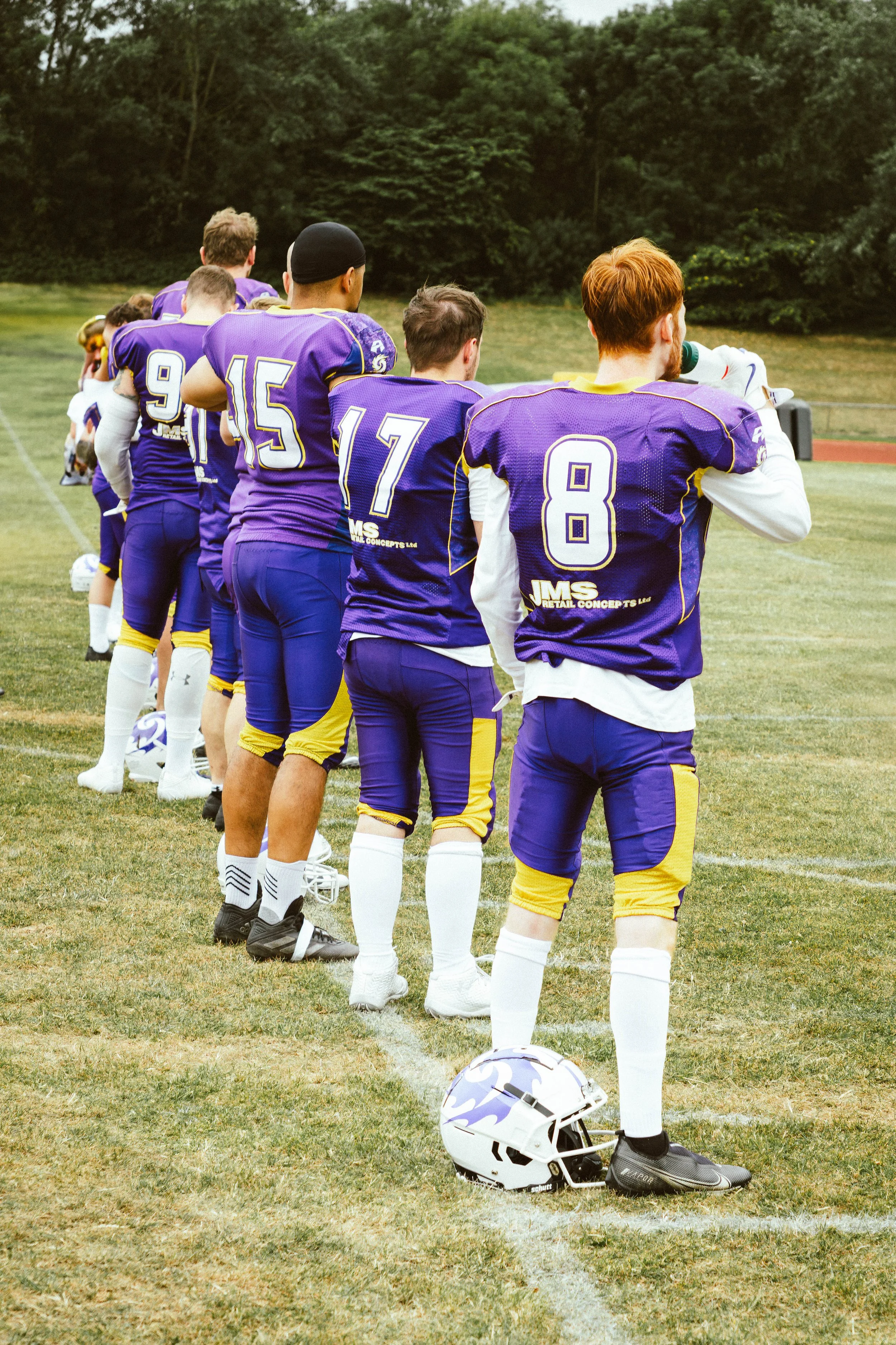 A group of football players in purple and yellow uniforms standing in a line on a field, with one player holding a microphone and wearing a helmet on the ground nearby.