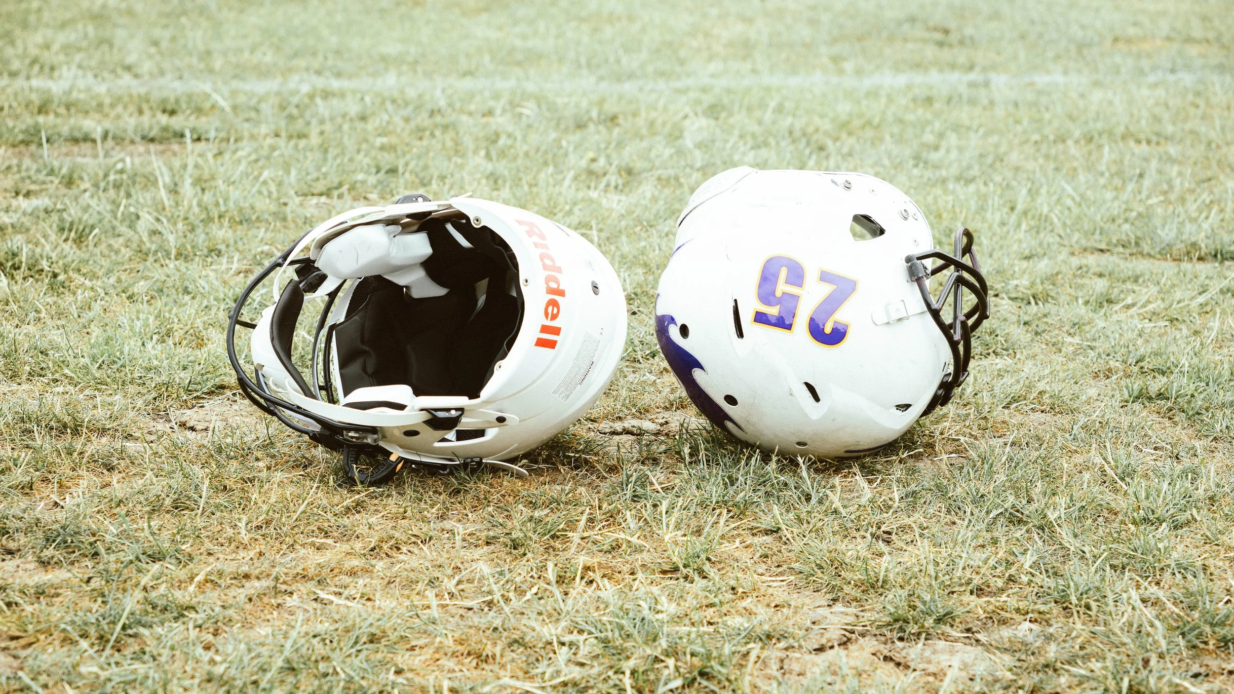Two American football helmets on grass field, one white with orange and black accents, the other white with purple and gold accents.