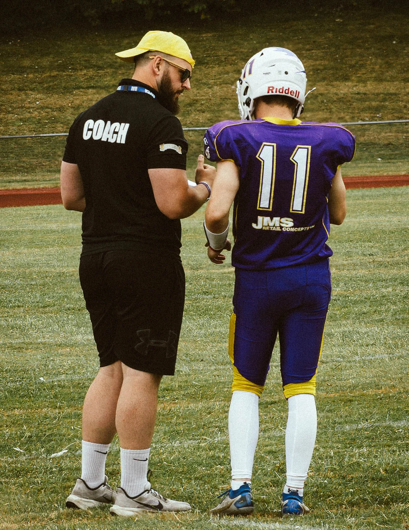 A football coach wearing black shorts and a black t-shirt with 'COACH' printed on the back, speaking to a football player in purple and yellow uniform with the number 11, on a grassy football field.
