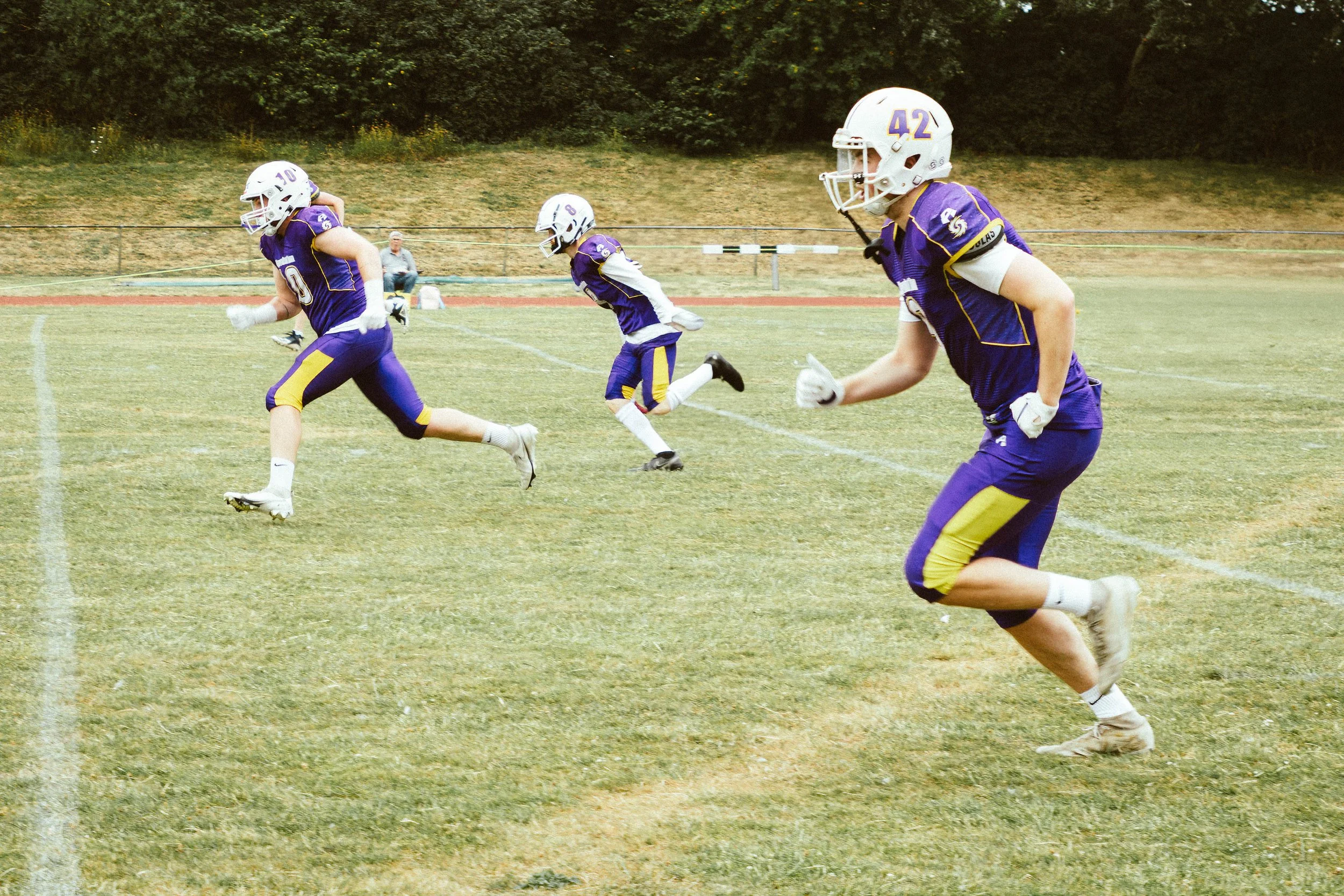 Youth football players in purple and yellow uniforms running on the field during a game.