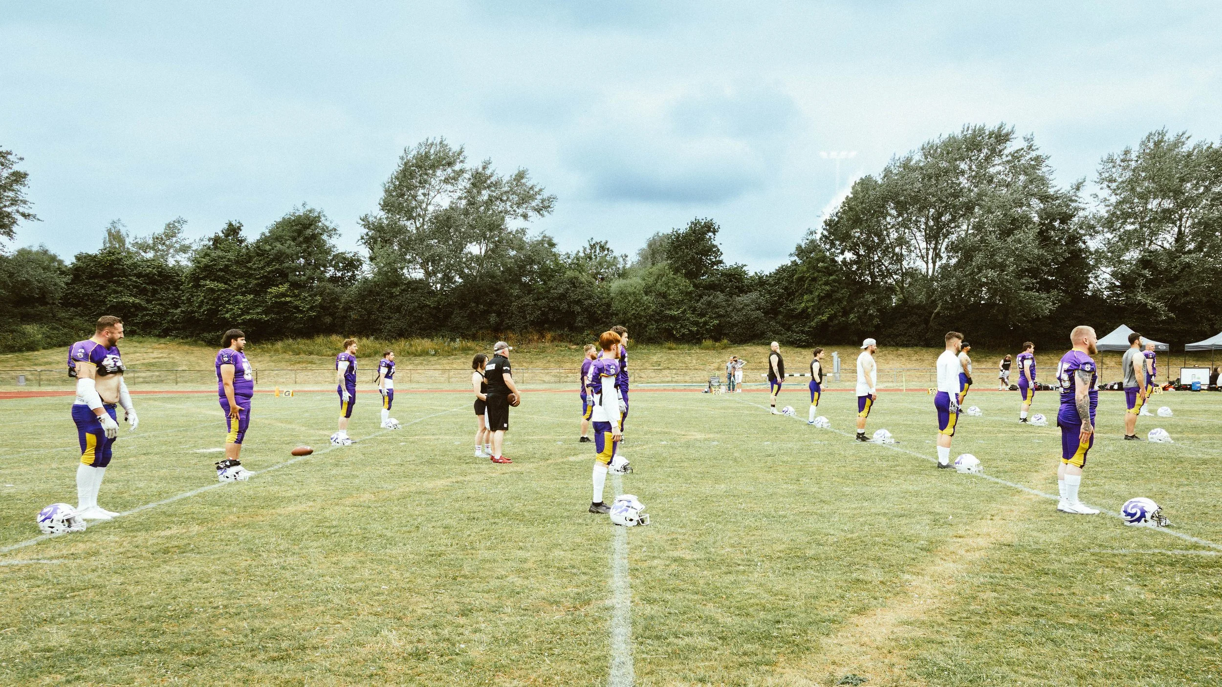 American football team practicing on a field with players standing in formation, wearing purple and yellow uniforms, helmets on the ground, and coaches observing.