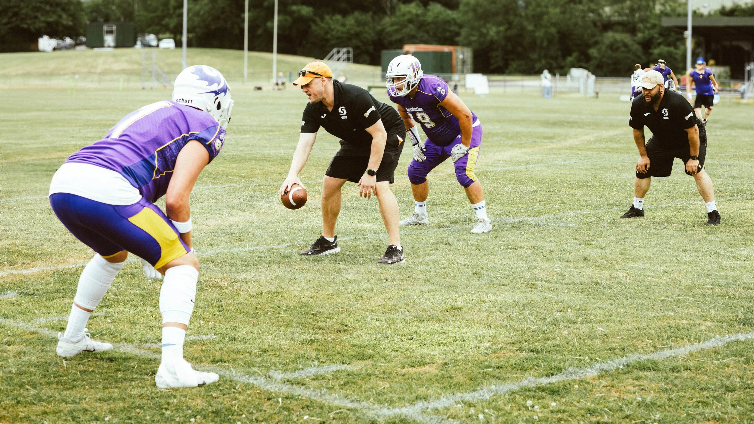 Football players in purple and black uniforms on a field during practice, with one player crouched and others in defensive positions