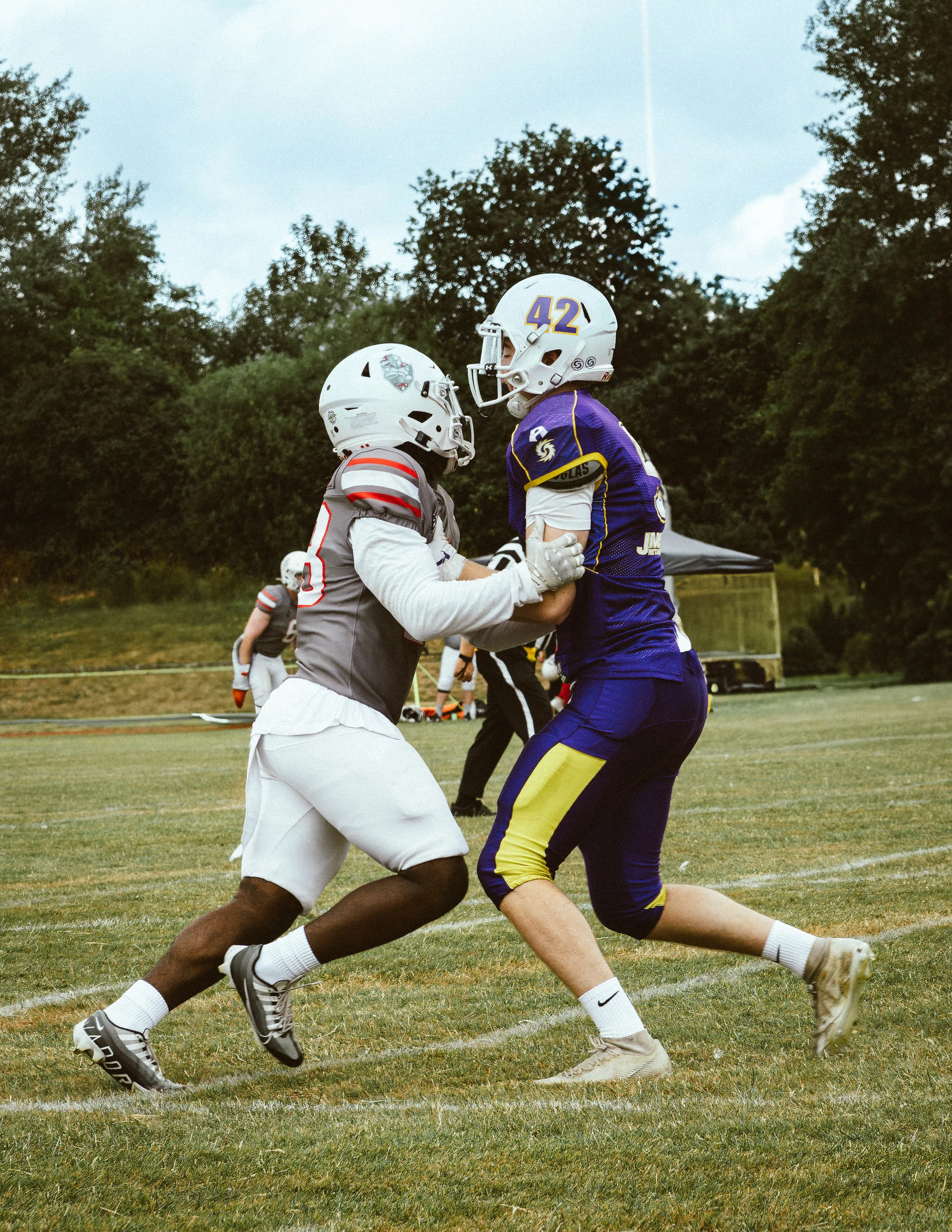 Two football players wearing helmets and uniforms are engaged in a tackle during a game on a grassy field, with trees and a tent in the background.