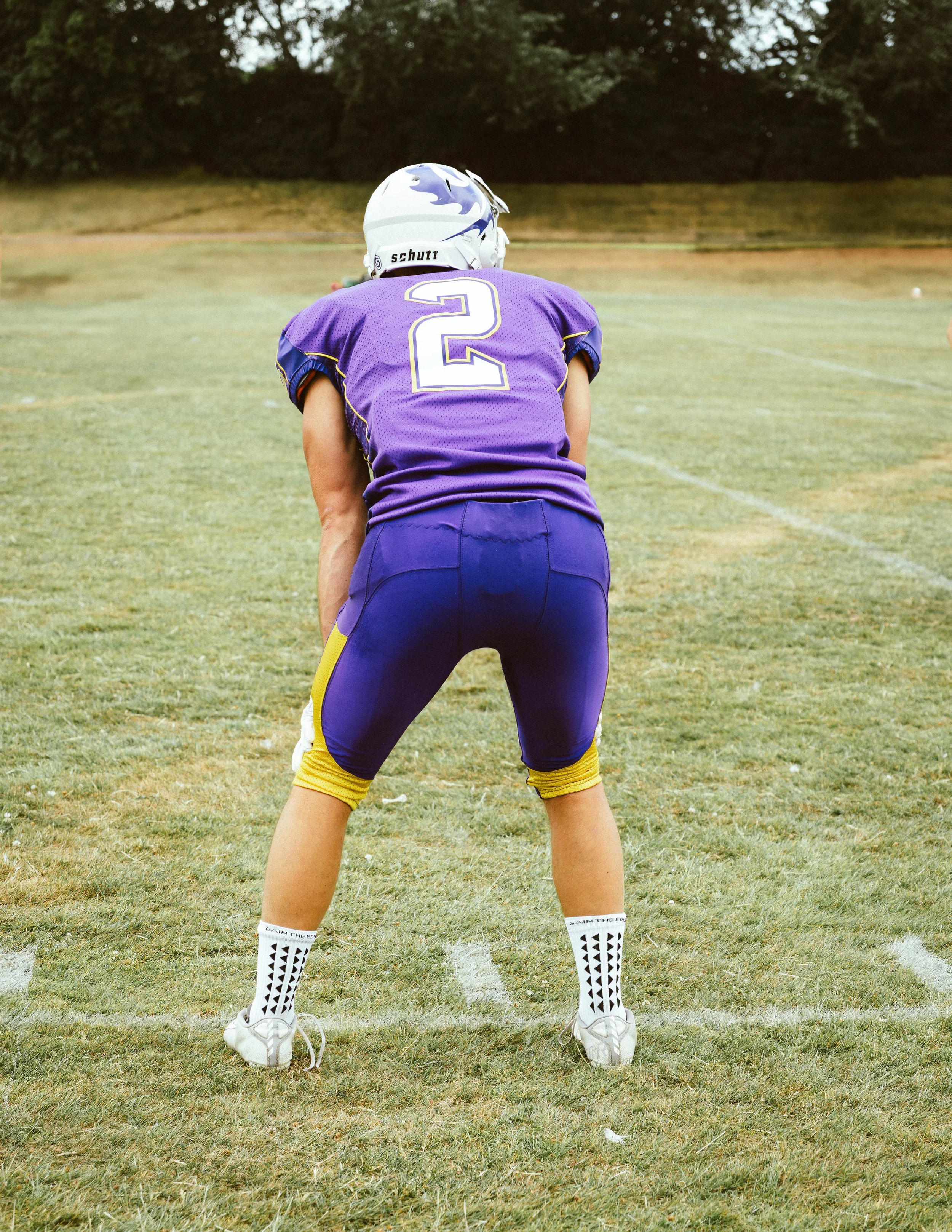 A football player in purple uniform with the number 2, wearing a helmet and white cleats, is seen from the back, standing on a grassy field.