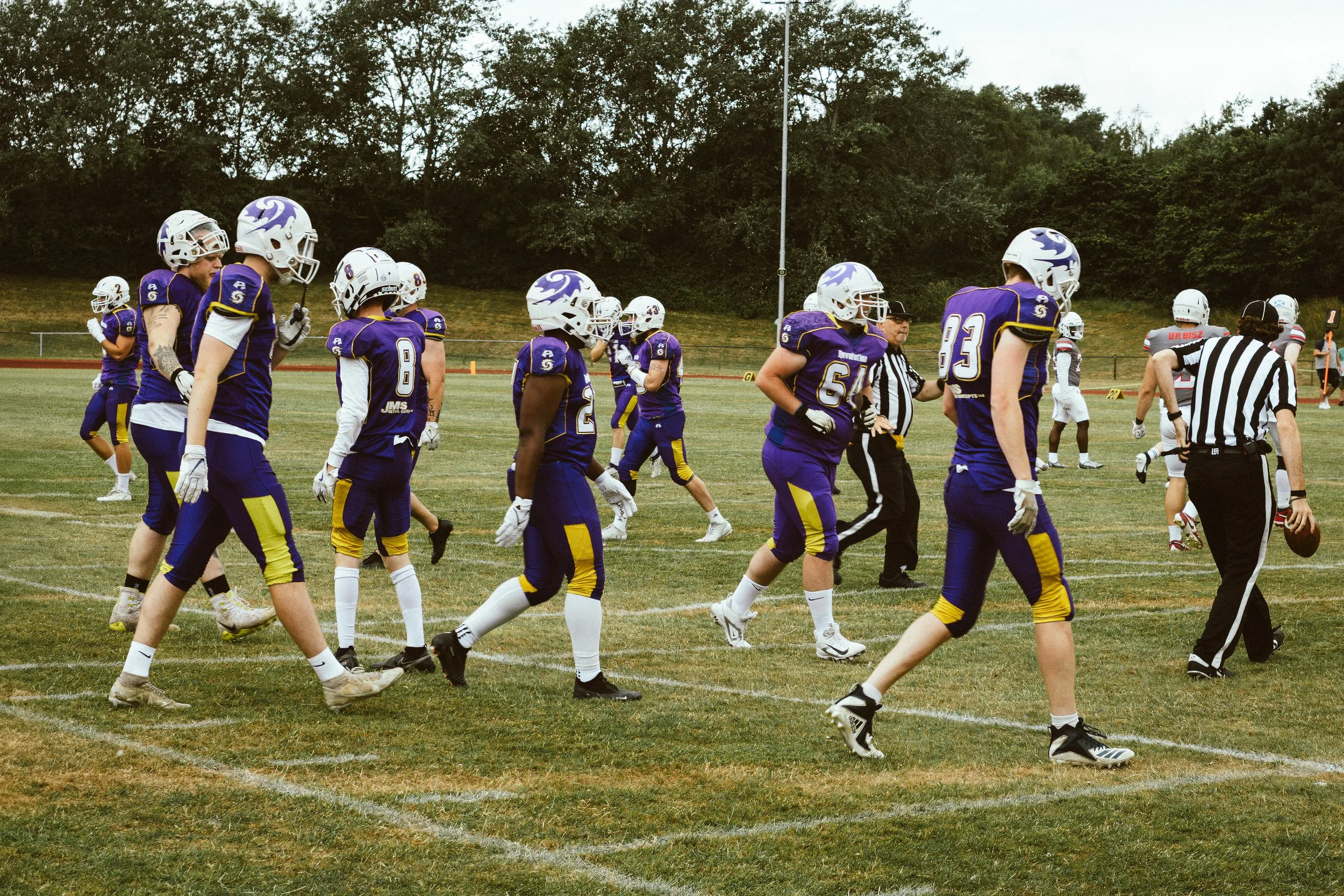 A group of young football players in purple and yellow uniforms walking on a football field, with referees and opposing players in the background.