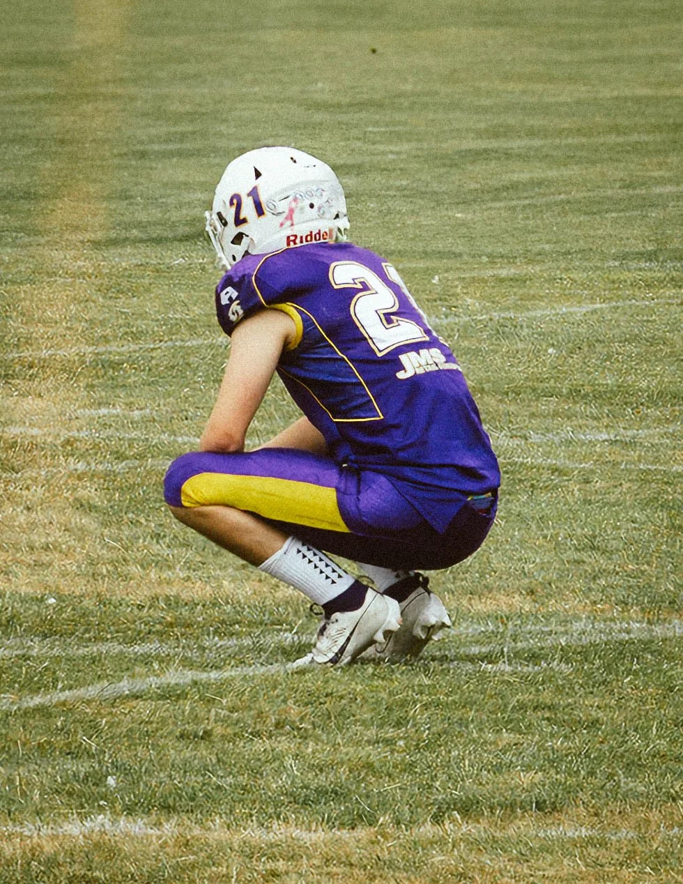 A young football player in purple and yellow uniform crouching on the field during a game, wearing a white helmet with the number 21 and a white faceguard.