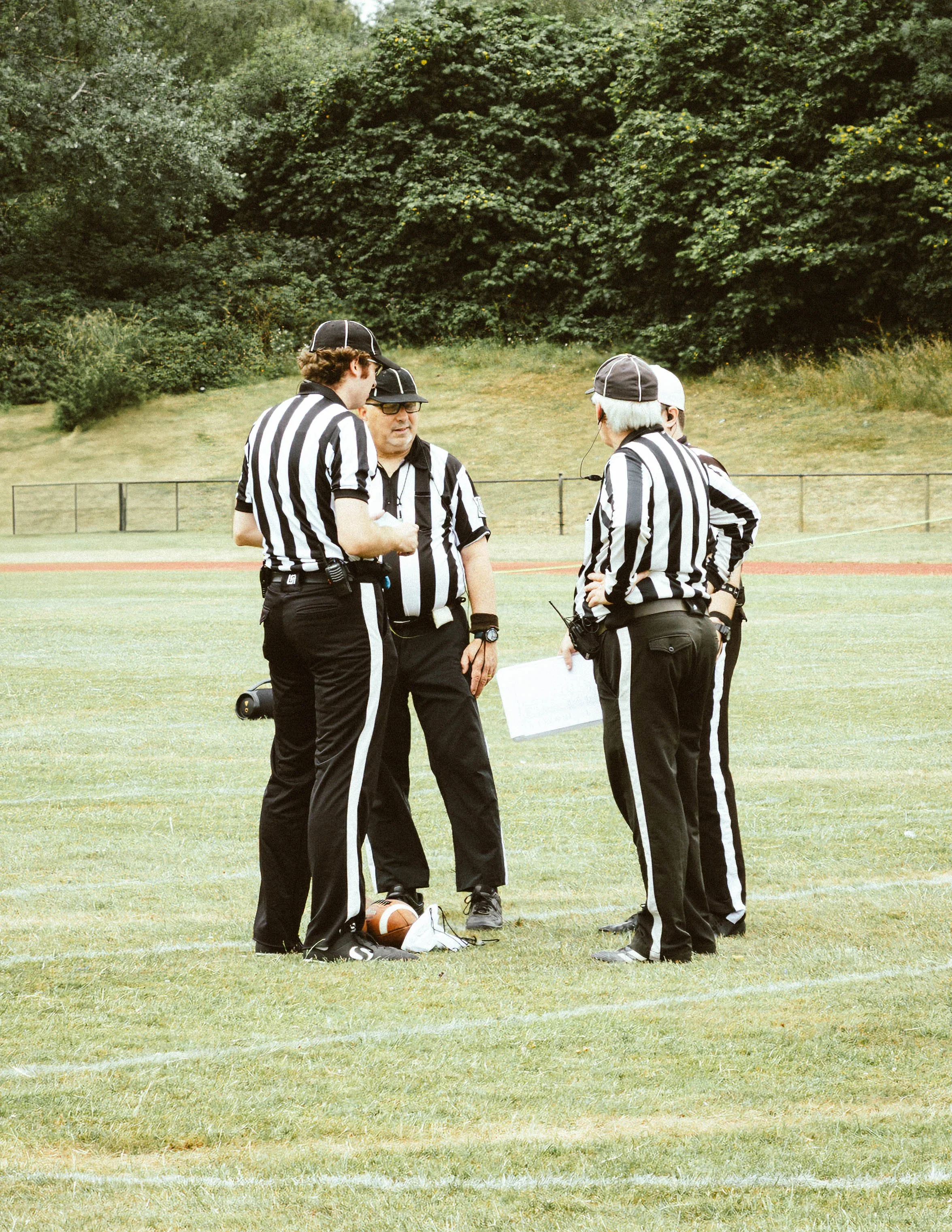 Group of referees in black and white striped shirts standing on a grassy field, discussing with each other during an outdoor sports event.