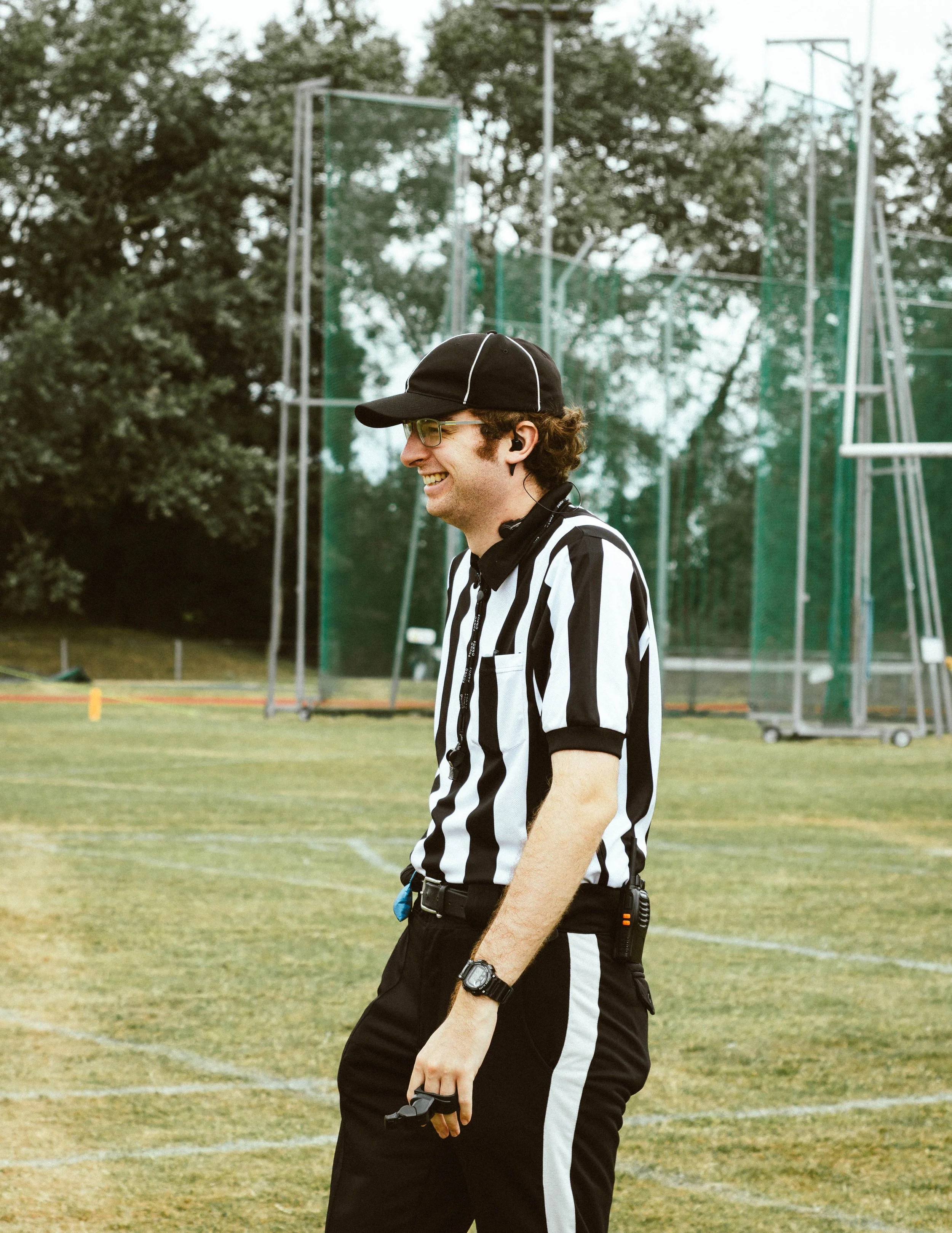 Male football referee smiling on a field, wearing a black cap, black and white striped referee shirt, black pants, and holding a whistle, with a scoreboard and trees in the background.