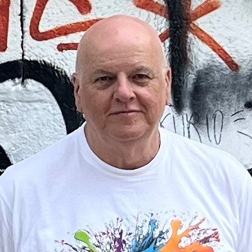 Andrew Bowers, artist wearing t-shirt with a colorful paint splash design standing in front of a graffiti wall.