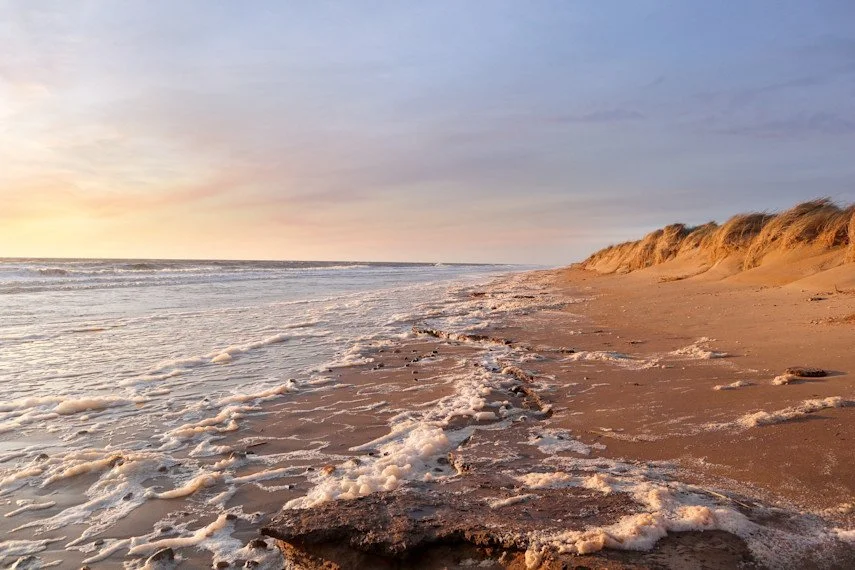 Beach at sunset with foam-covered waves and sandy dunes.