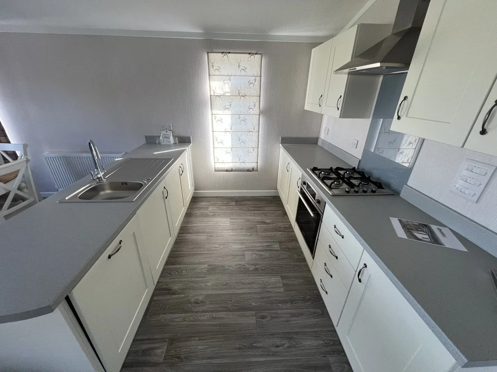 Empty kitchen with white cabinets, gray countertops, a window with a floral curtain, a stove, oven, and a stainless steel sink.