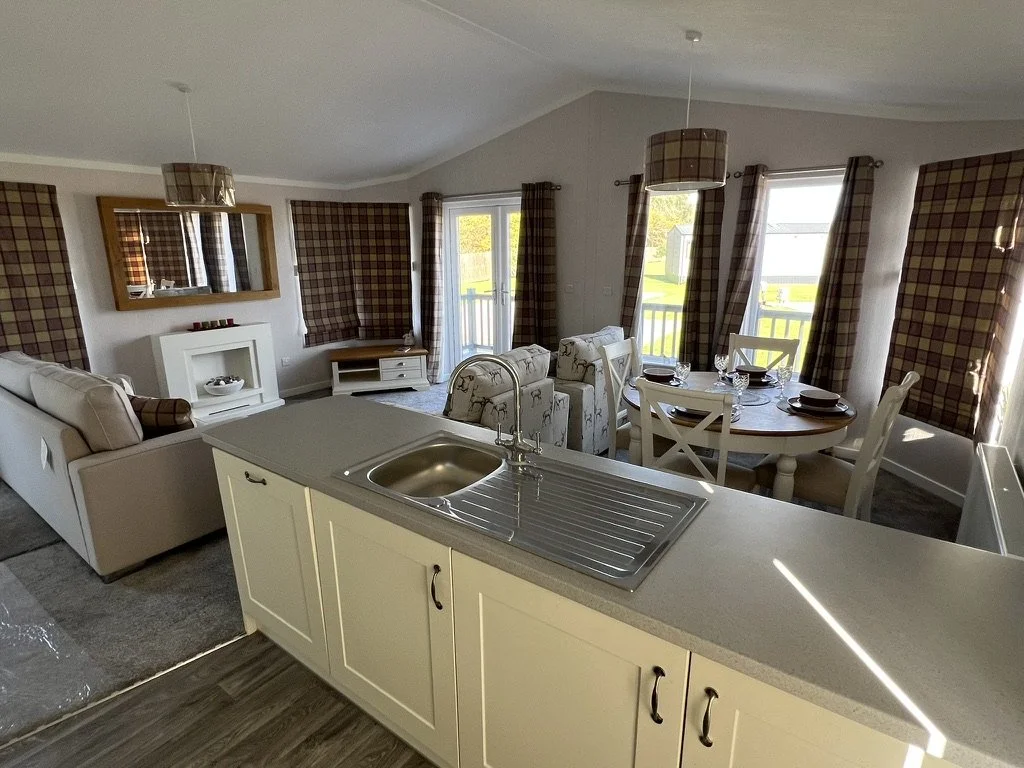 Open-concept living room and dining area with a kitchen island, beige sofas, and a round dining table set for four, with windows and plaid curtains.