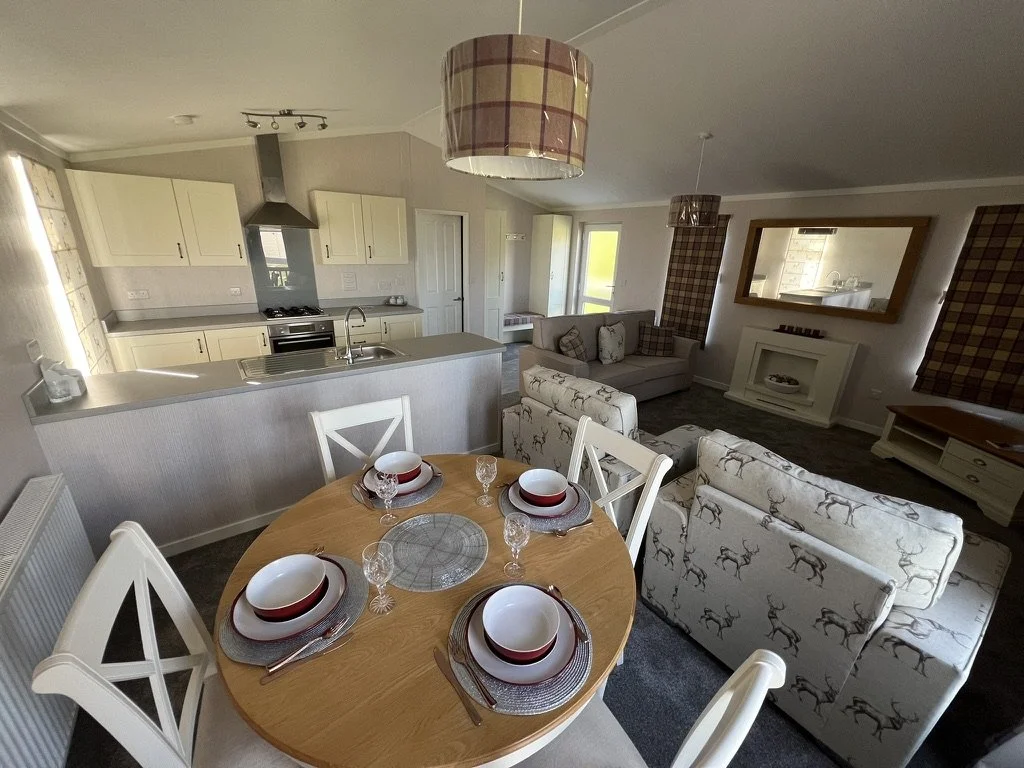 Open-concept kitchen and living room with a round dining table set with four place settings, a kitchen with cream cabinets, a gray countertop, and a living area with patterned sofas, a mirror, and a fireplace.