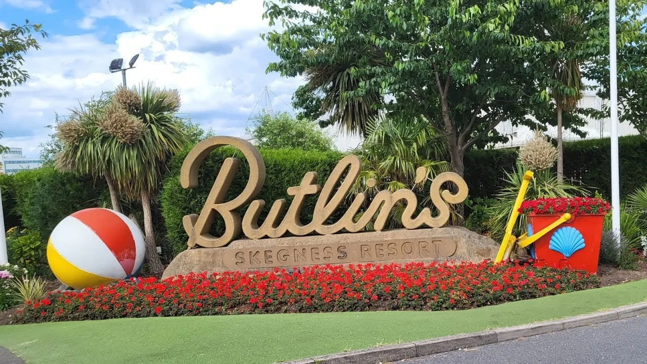 Sign reading "Butlin's Skegness Resort" with a beach ball, red flower planter, and yellow beach toy on green grass, surrounded by trees and bushes.