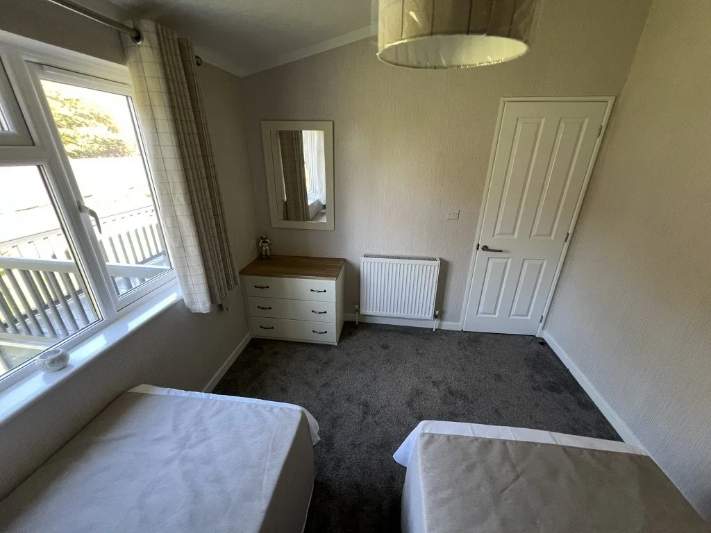 A small bedroom with two single beds, a window with checkered curtains, a white dresser with a wooden top, a mirror on the wall, a radiator, and a closed white door. The ceiling has a lampshade.