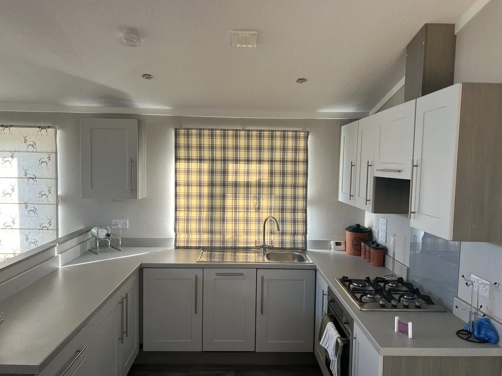 Kitchen with white cabinets, a sink beneath a window with a plaid curtain, a gas stove, and counter space with some containers and a small dish drying rack.