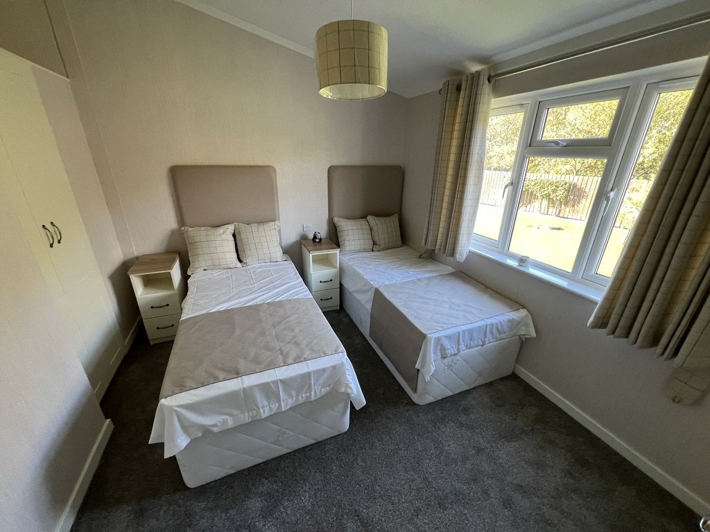 A bedroom with two single beds, each with beige headboards, white bedding, and beige pillows. There are small white nightstands beside each bed, one with a clock. A large window with beige curtains looks outside to a green yard. The room has cream-co