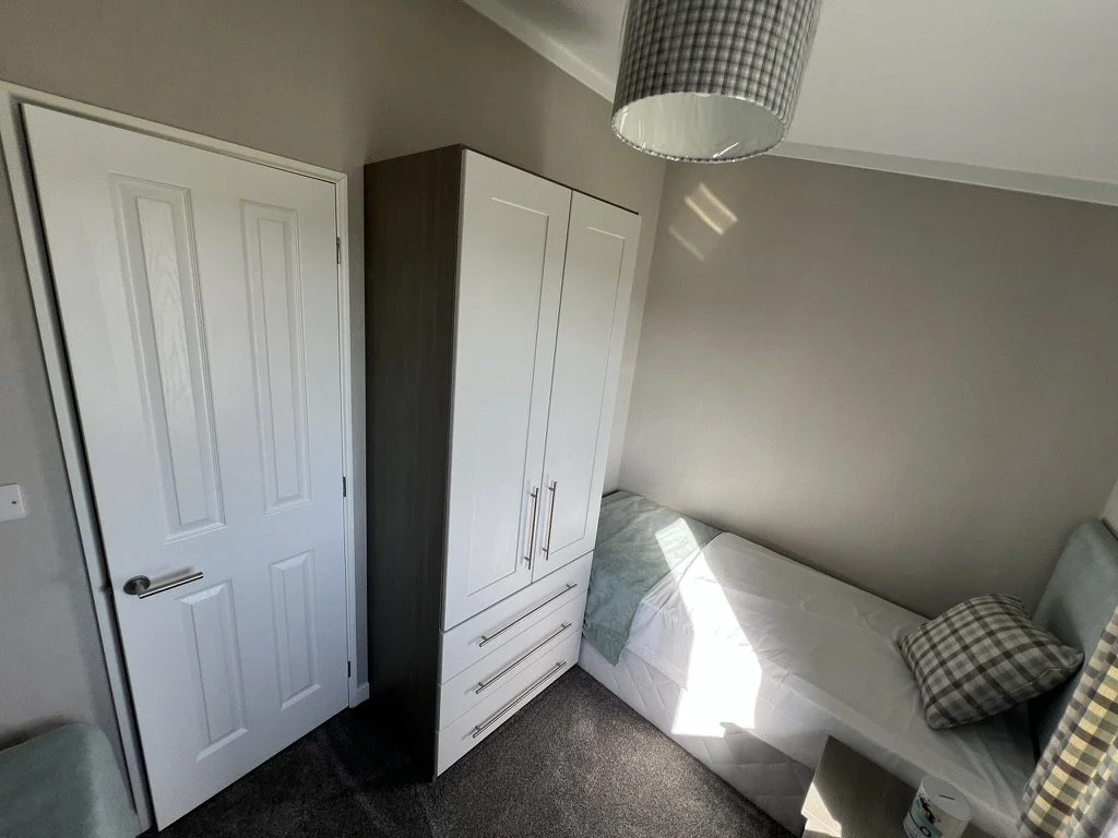 A small bedroom with a bed, a wardrobe, a door, a hanging ceiling light, a pillow, and some sunlight streaming through a window.