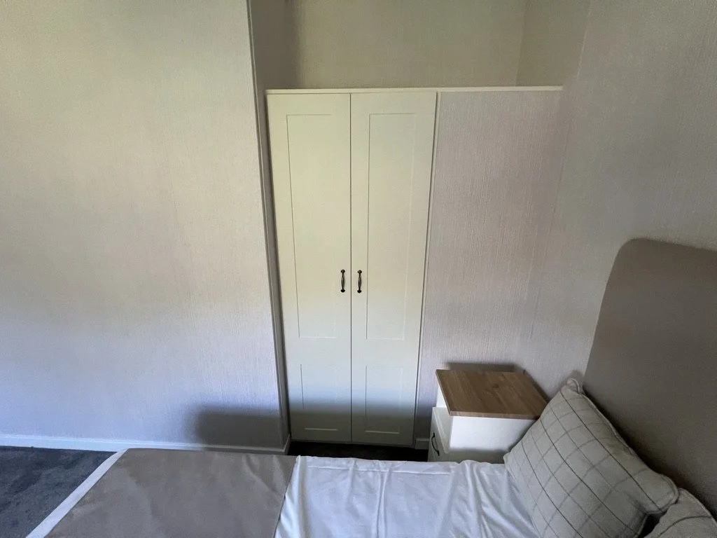 A bedroom corner with a white wardrobe, small nightstand with a wooden top, bed with a pillow, and a gray headboard.