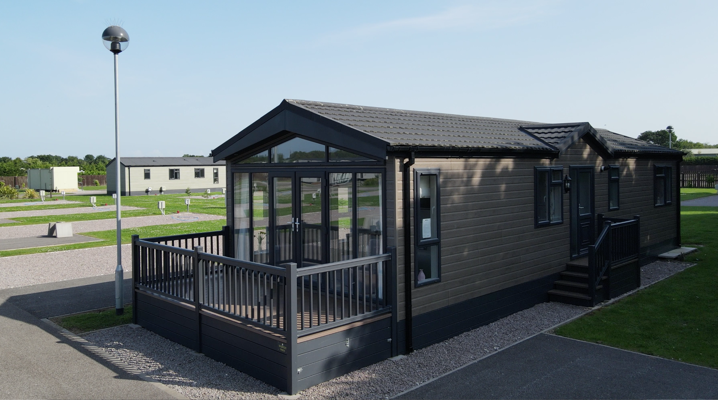 A modern tiny house holiday lodge with a wooden exterior, black railings, and large windows, situated in a well-maintained community area.