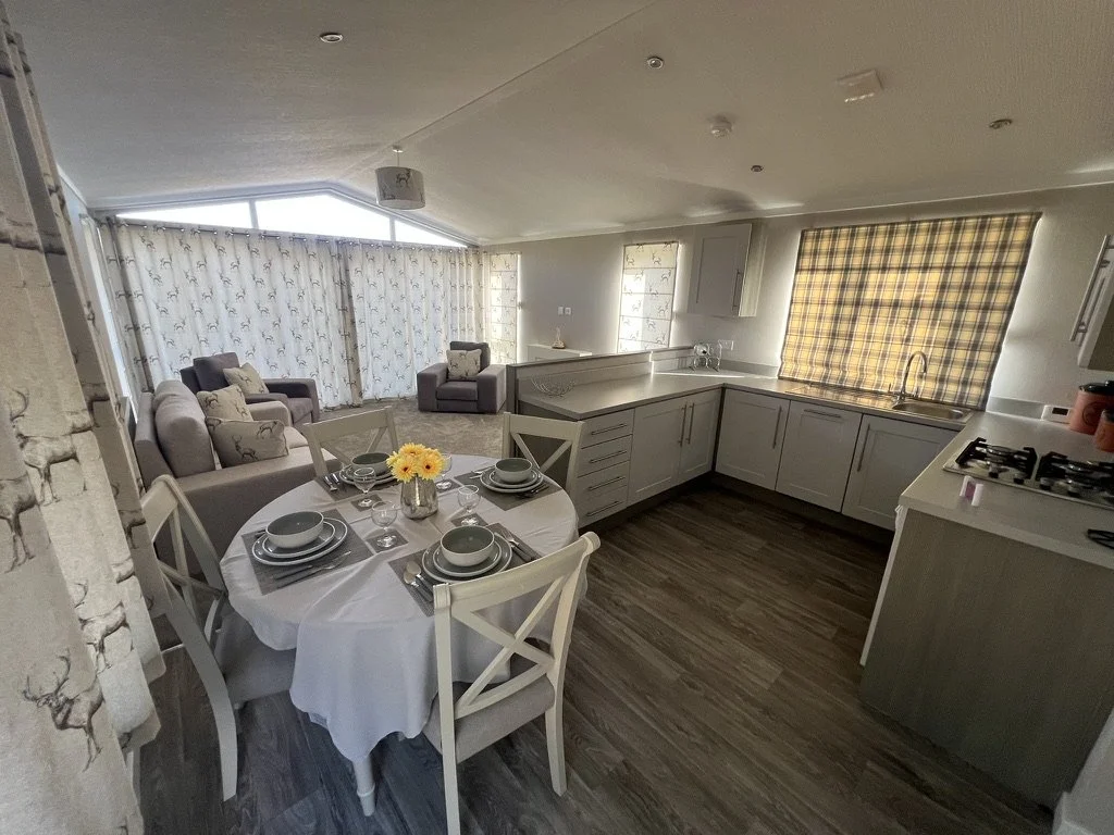 Open-concept kitchen and living room with dining table set, sitting area with four armchairs, large windows with curtains, and a kitchen with white cabinets, stove, and sink.