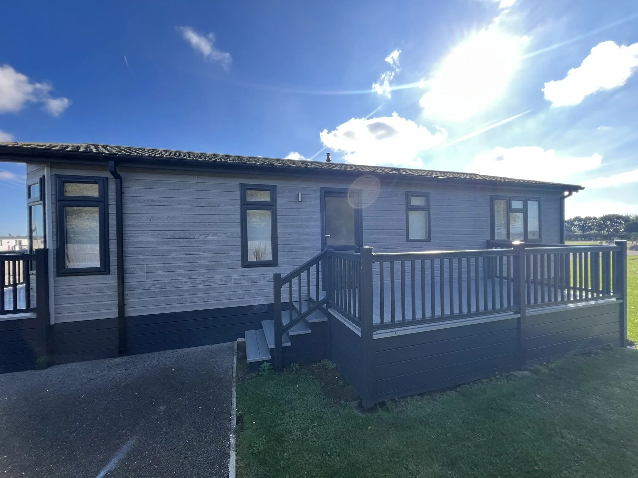 A lodge exterior with light grey horizontal cladding, grey window frames, a porch with stairs, and a black fence. The sky is clear with some clouds and the sun bright.
