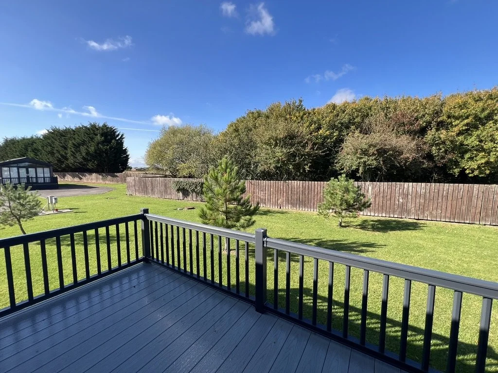 View of a backyard with a wooden deck, green grass, small trees, a fence, and a clear blue sky with a few clouds.