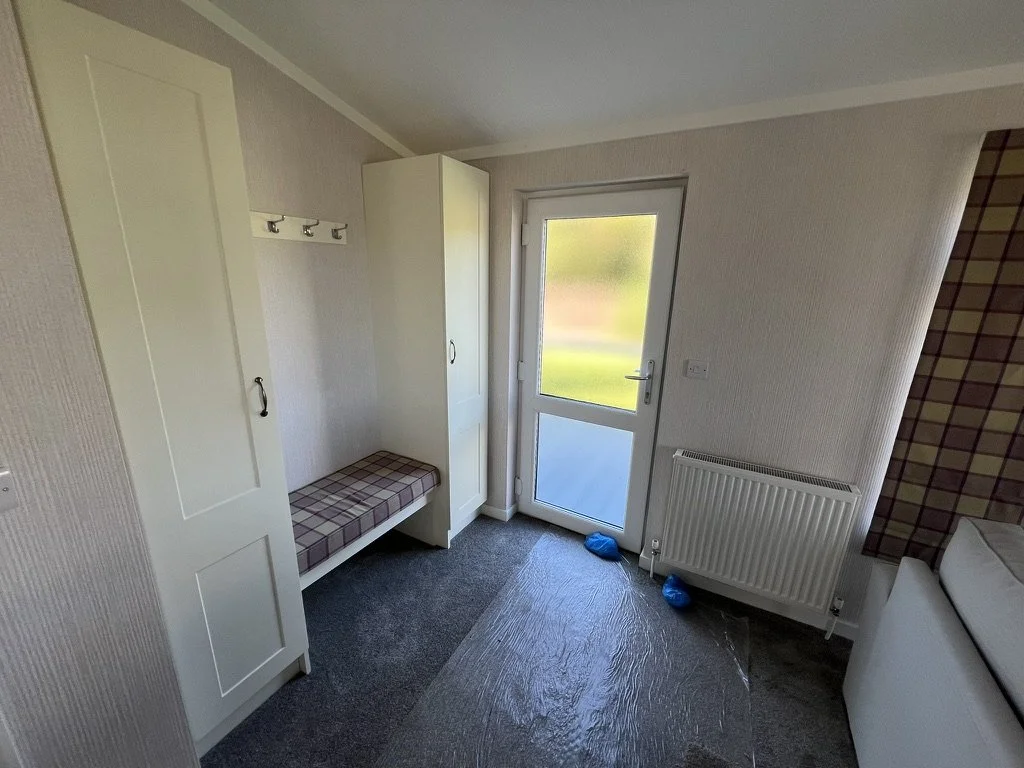 Small room with white door, wardrobe, coat hooks, bench with plaid cushion, radiator, and partial view of bed. Green outdoor scene visible through glass door.