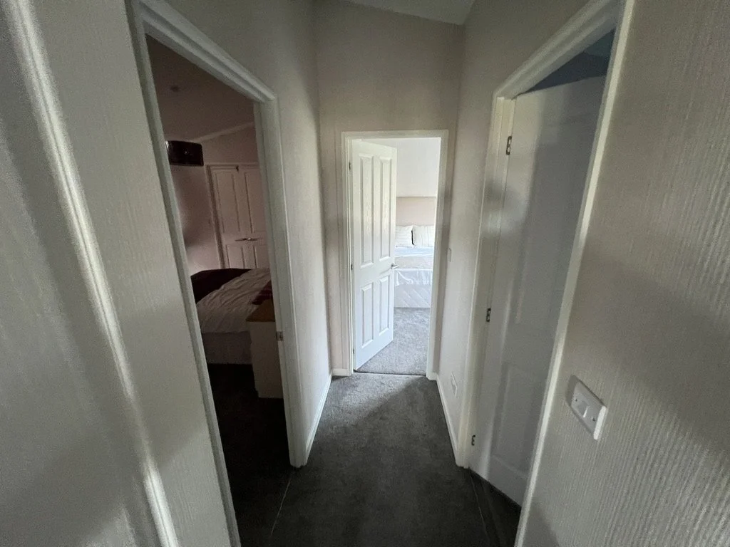 Hallway view with three open doors leading to bedrooms, with beige textured walls and gray carpet flooring.