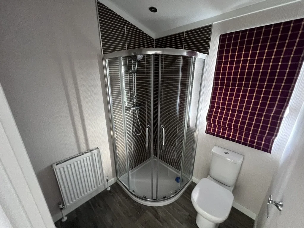 Small bathroom featuring a corner shower with glass doors, a toilet, a maroon and yellow checkered window blind, a radiator on the left wall, and wood-like flooring.