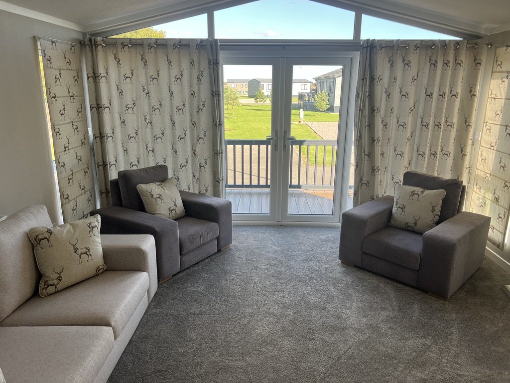 Living room with three armchairs and two sofas, all with furniture cushions featuring reindeer designs, and large windows with matching reindeer curtains showing a green yard outside.
