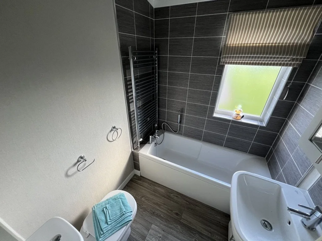 Bathroom with a bathtub, window, sink, and toilet, featuring dark gray tiled walls and a beige painted wall.