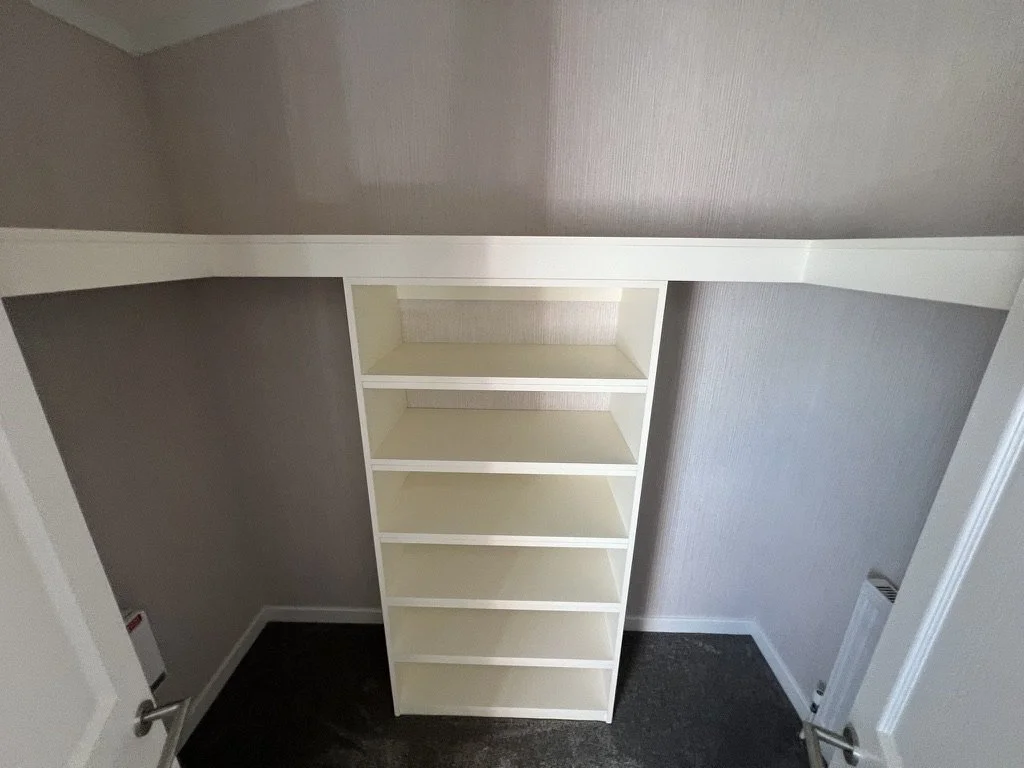 Empty white bookshelf with six shelves in a small hallway closet with beige walls and dark carpeted floor.