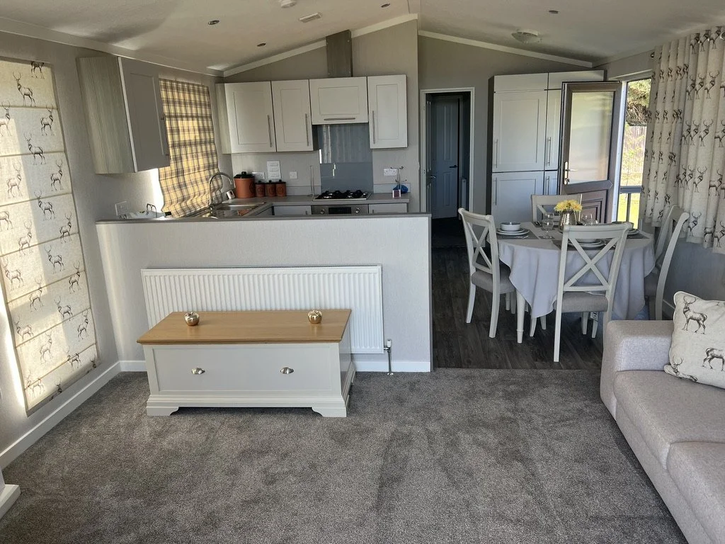 Living room and kitchen area with a beige sofa, a white table, and white kitchen cabinets, with sunlight coming through curtains on windows.