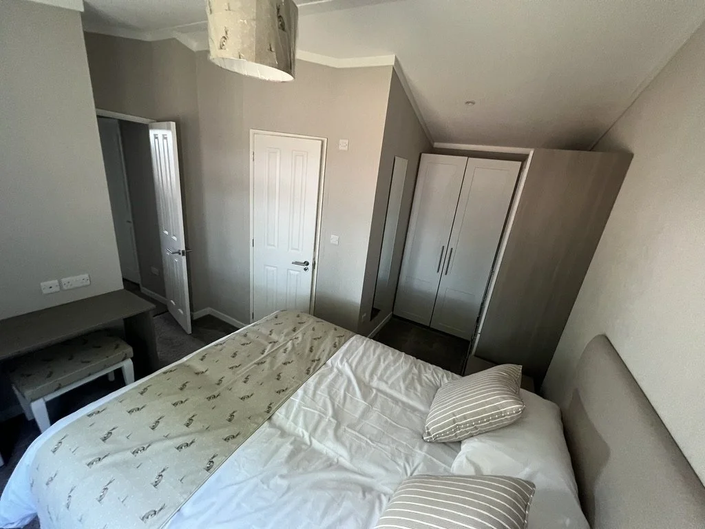 A bedroom with a bed covered in white sheets and striped pillows, a small desk with a stool, a built-in wardrobe, and a door leading to another room or closet, with a hanging light fixture above.
