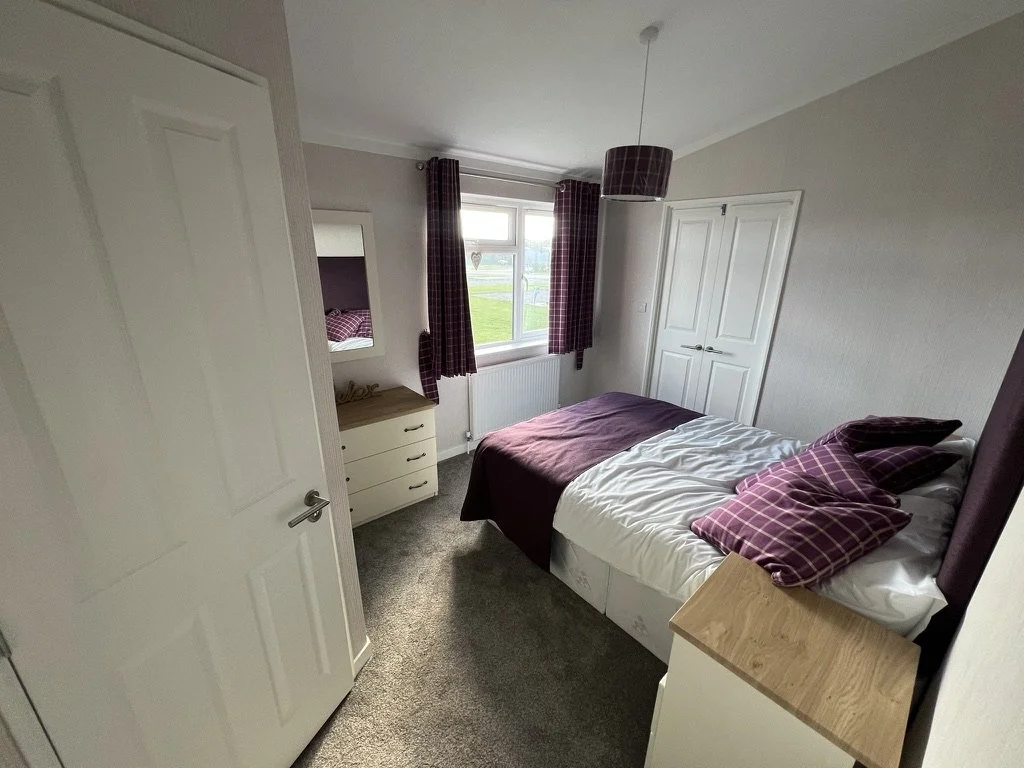 A small bedroom with a bed, pillows, and a window with curtains. There is a wardrobe and a dresser, with a mirror on the wall.