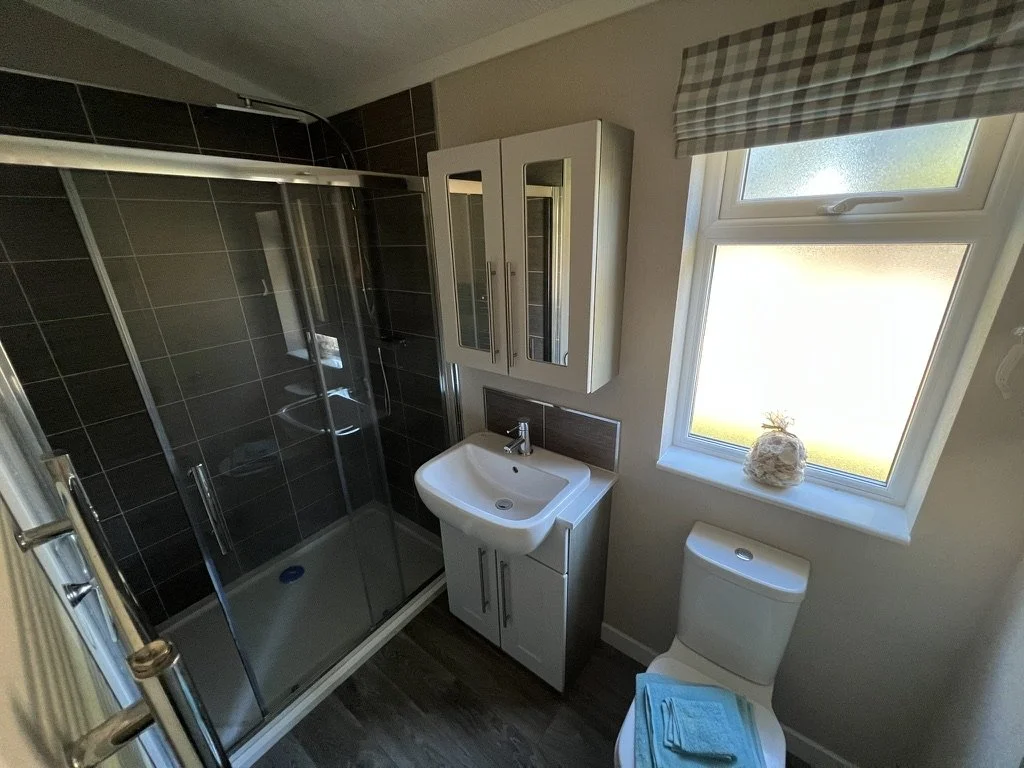 Small bathroom with a glass shower enclosure, a white sink with storage below, a wall-mounted mirrored cabinet, a toilet with a turquoise towel on top, and a window with a checkered curtain letting in sunlight.