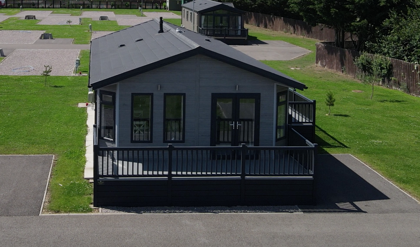 A small modern house with a gray exterior and black trim, featuring a deck with black railings, surrounded by green grass and small trees, with a parking area in the foreground.