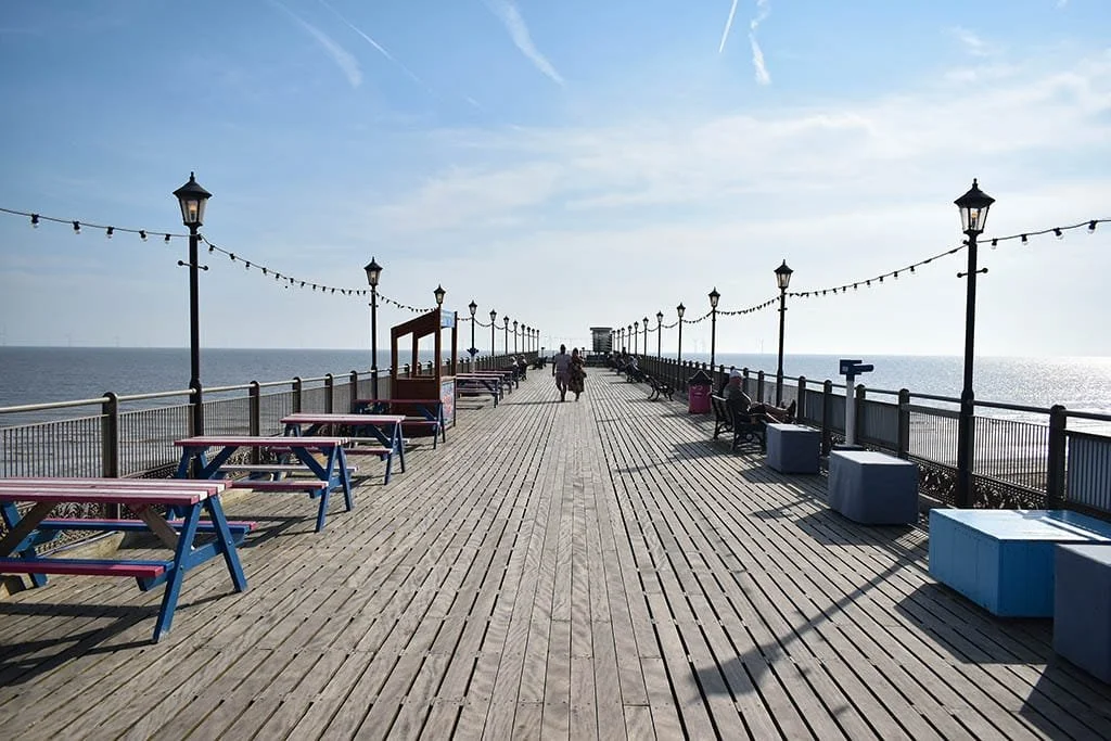 A wooden pier extending over the ocean with benches, street lamps, and string lights, with a few people walking and sitting, under a blue sky with some clouds.