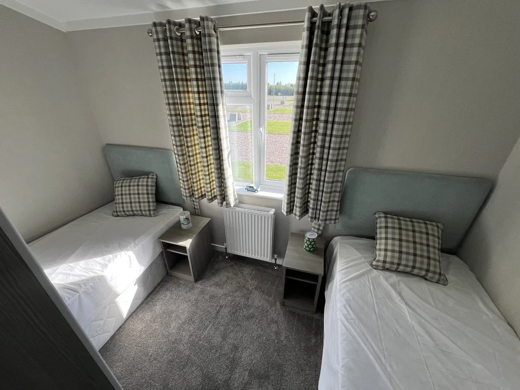 A small bedroom with two single beds, each with a plaid pillow and headboard, between them a nightstand with a lamp and mug, behind the beds a window with plaid curtains showing a view of a grassy area outside.